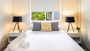 Una cama bien hecha con edredón blanco, dos mesitas de noche con lámparas, una ventana con vistas a zonas verdes y un aire acondicionado instalado en la pared.