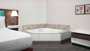 Una habitación de hotel moderna con una cama grande, un baño separado con bañera y una mesita de noche pequeña con lámpara.