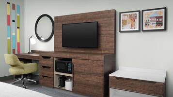 Habitación de hotel moderna con una unidad de entretenimiento de madera, televisor de pantalla plana, escritorio con silla, mesita de noche blanca y un espejo grande.