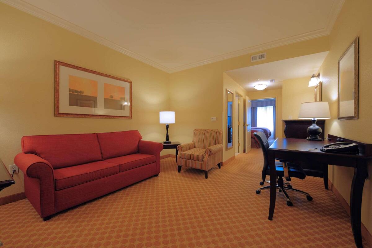 A hotel room with a red sofa, a striped armchair, a desk with a lamp, and a framed picture on the wall.