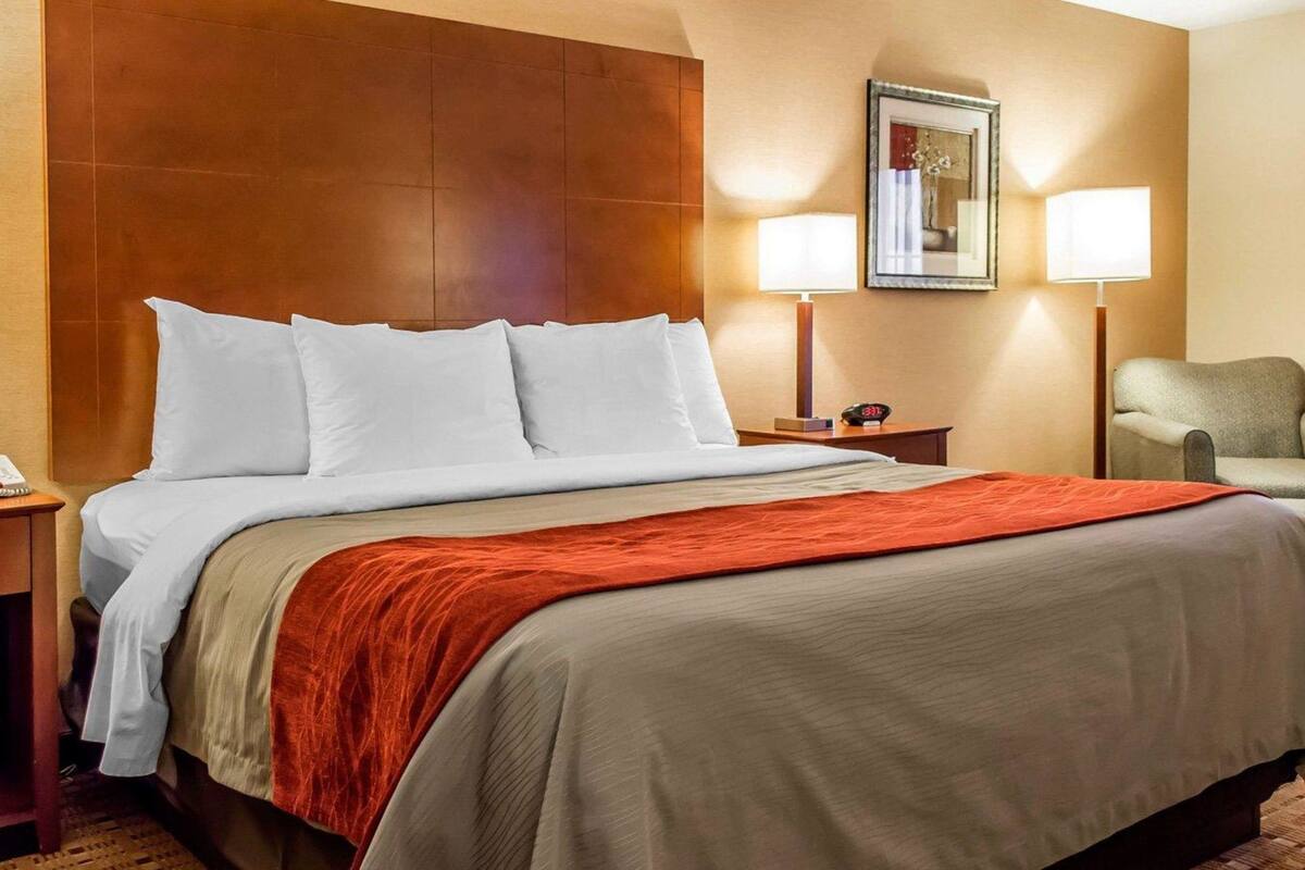 Une chambre d’hôtel avec un grand lit, une tête de lit en bois, deux lampes de chevet, une chaise et un tableau encadré au mur.