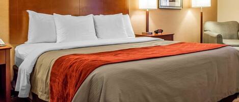 A hotel room with a large bed, a wooden headboard, two bedside lamps, a chair, and a framed picture on the wall.