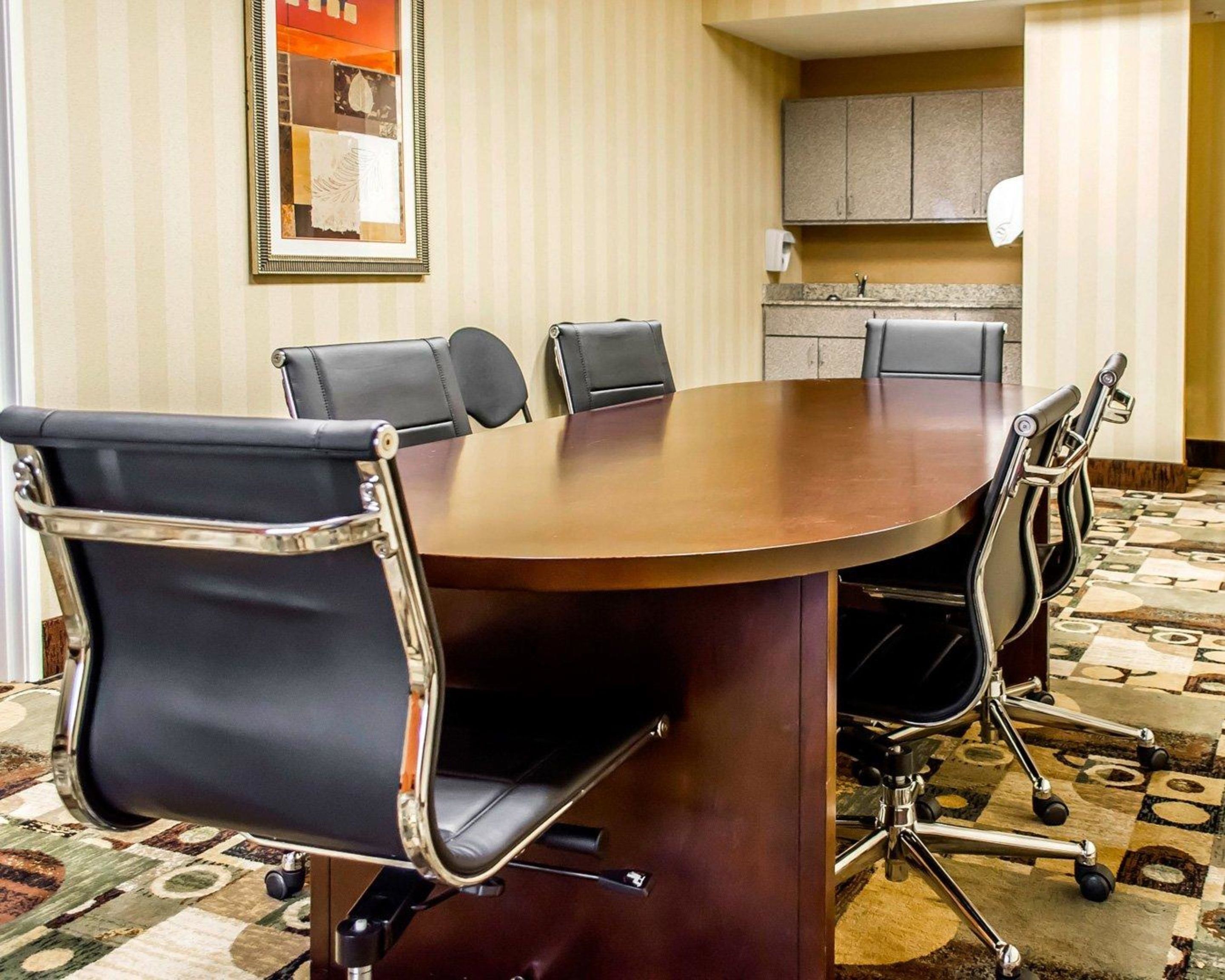 A conference room with a round table, six black leather chairs with chrome frames, a patterned carpet, a framed artwork on the wall, and a kitchenette in the background.
