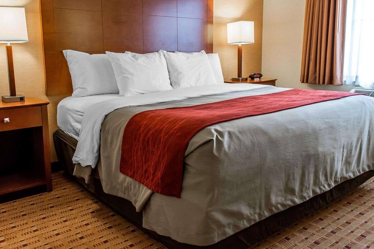 Une chambre d’hôtel avec un grand lit, deux tables de chevet, une petite table, un plaid rouge et une fenêtre avec des rideaux.