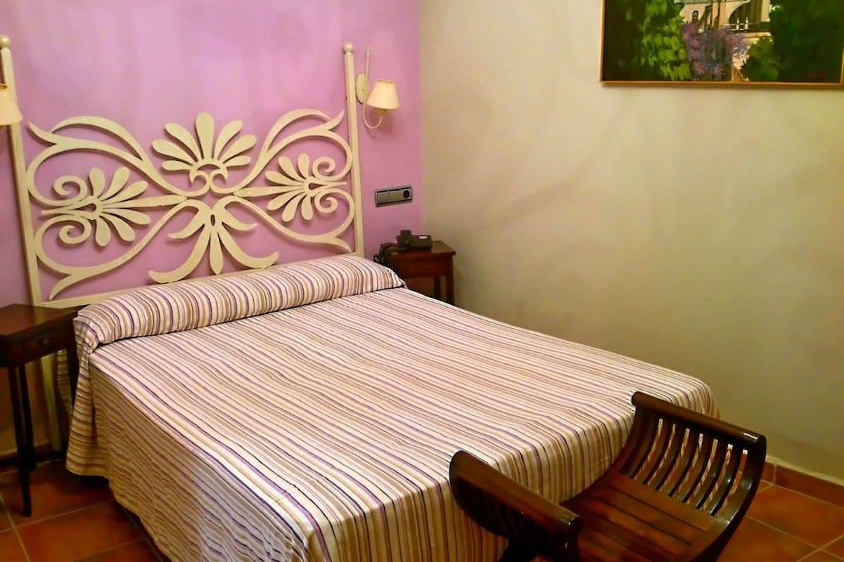 Un dormitorio con cama, una silla, una mesita de noche y una lámpara de pared.