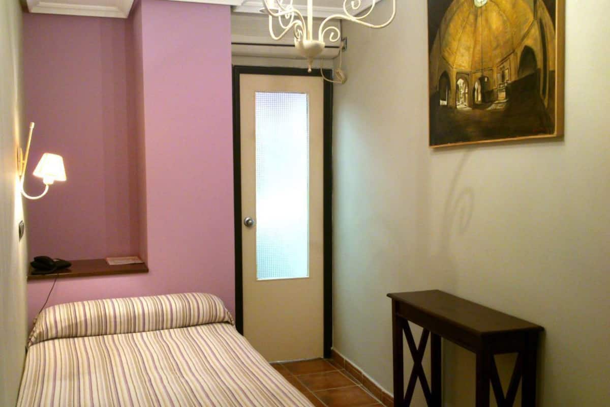 Un dormitorio con una cama, una mesita, un cuadro, una puerta y una ventana.