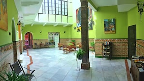 A spacious lobby with a high ceiling, tiled floor, and green walls. There are several seating areas with chairs and tables, and a bookshelf with books. - Hotel Al Andalus Jerez (Jerez de la Frontera)