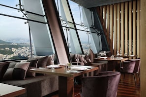 Un restaurante moderno con mesas altas y asientos acolchados, grandes ventanales con vistas a la ciudad.