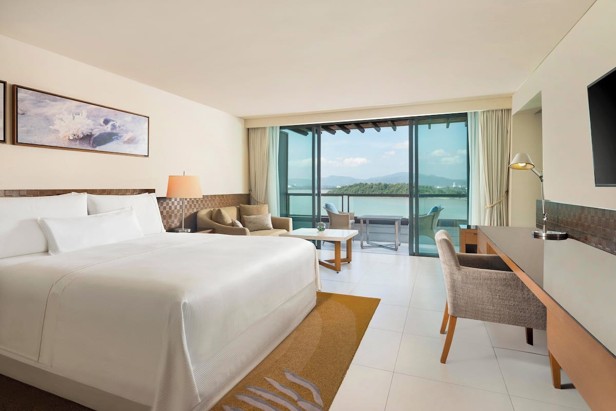 Une chambre d’hôtel avec un grand lit, un bureau, une chaise et une vue sur une étendue d’eau grâce à de grandes fenêtres.