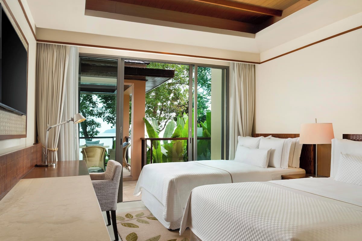 Une chambre d’hôtel avec deux lits, un bureau, une chaise, une lampe et une vue sur la verdure, accessible par une porte coulissante.