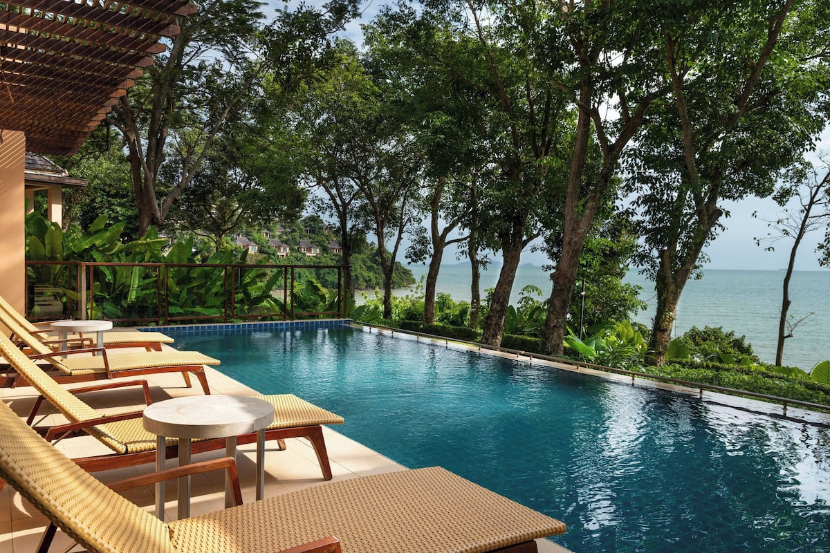 Un espace aménagé au bord de la piscine, avec des chaises longues et une petite table, entouré d’une végétation luxuriante et offrant une vue sur une étendue d’eau.
