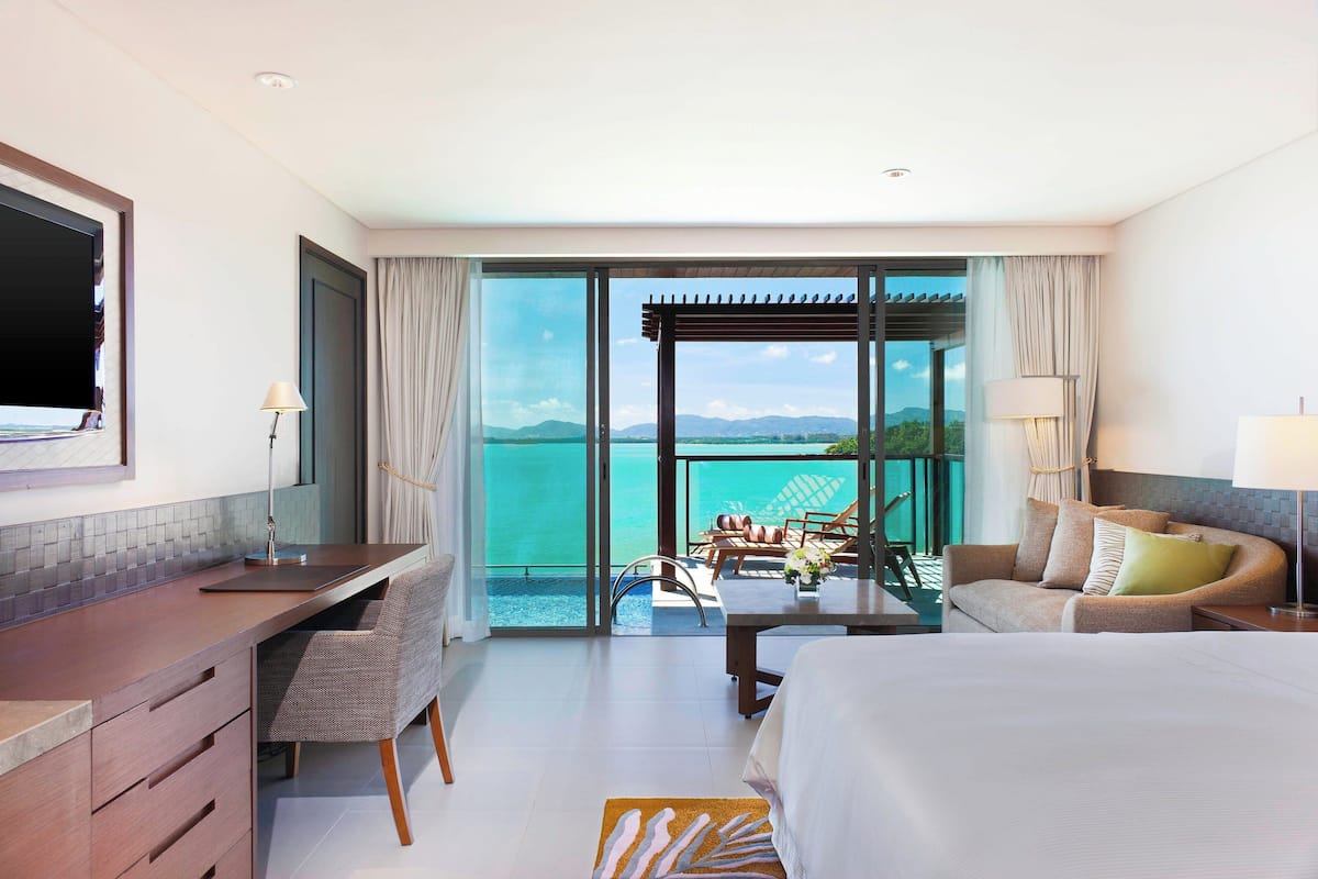 Une chambre d’hôtel avec un grand lit, un bureau avec une chaise, un balcon avec une table et des chaises, et une vue sur la mer.