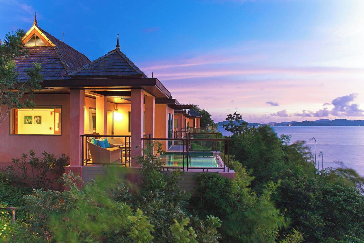 Une maison moderne dotée d’un balcon donnant sur une piscine, le tout entouré d’une végétation luxuriante.