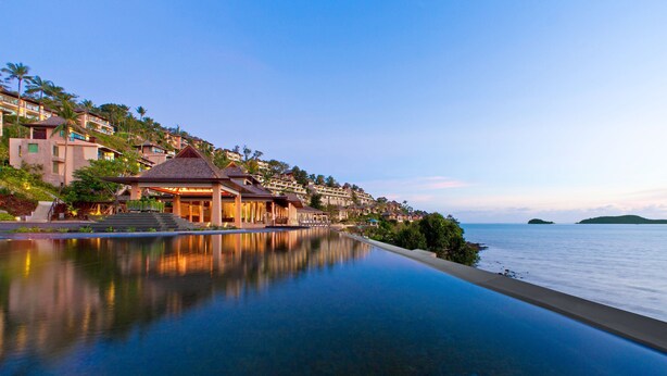 A resort with a long, reflective infinity pool, a covered seating area, and a view of the ocean and distant hills.