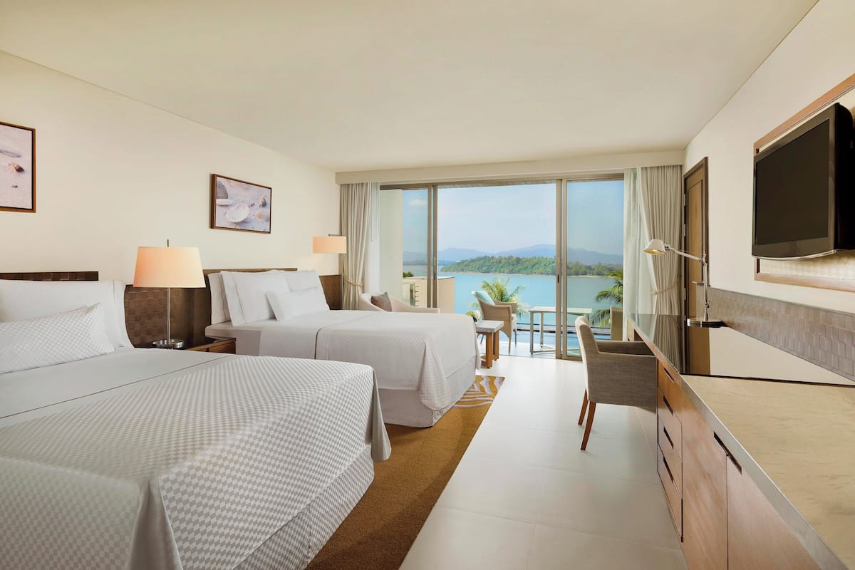 Une chambre d’hôtel avec deux lits, une télévision, un balcon donnant sur la mer et un bureau avec une chaise.