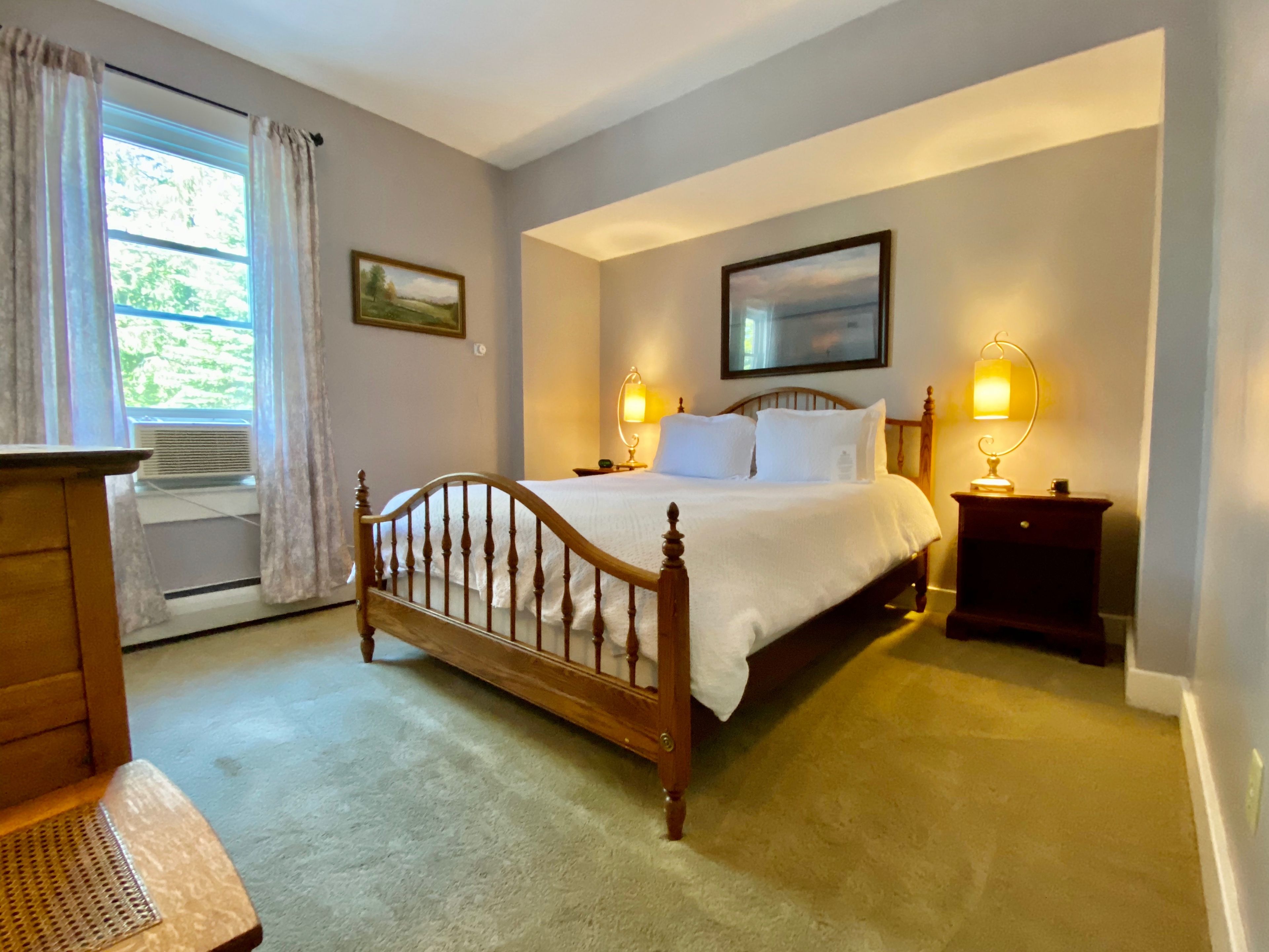 A bedroom with a wooden bed, two bedside tables with lamps, a window with curtains, and a framed painting on the wall.
