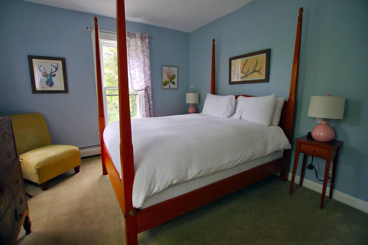 Un lit à baldaquin avec du linge de lit blanc, une table de chevet en bois avec une lampe, un fauteuil jaune et une commode.