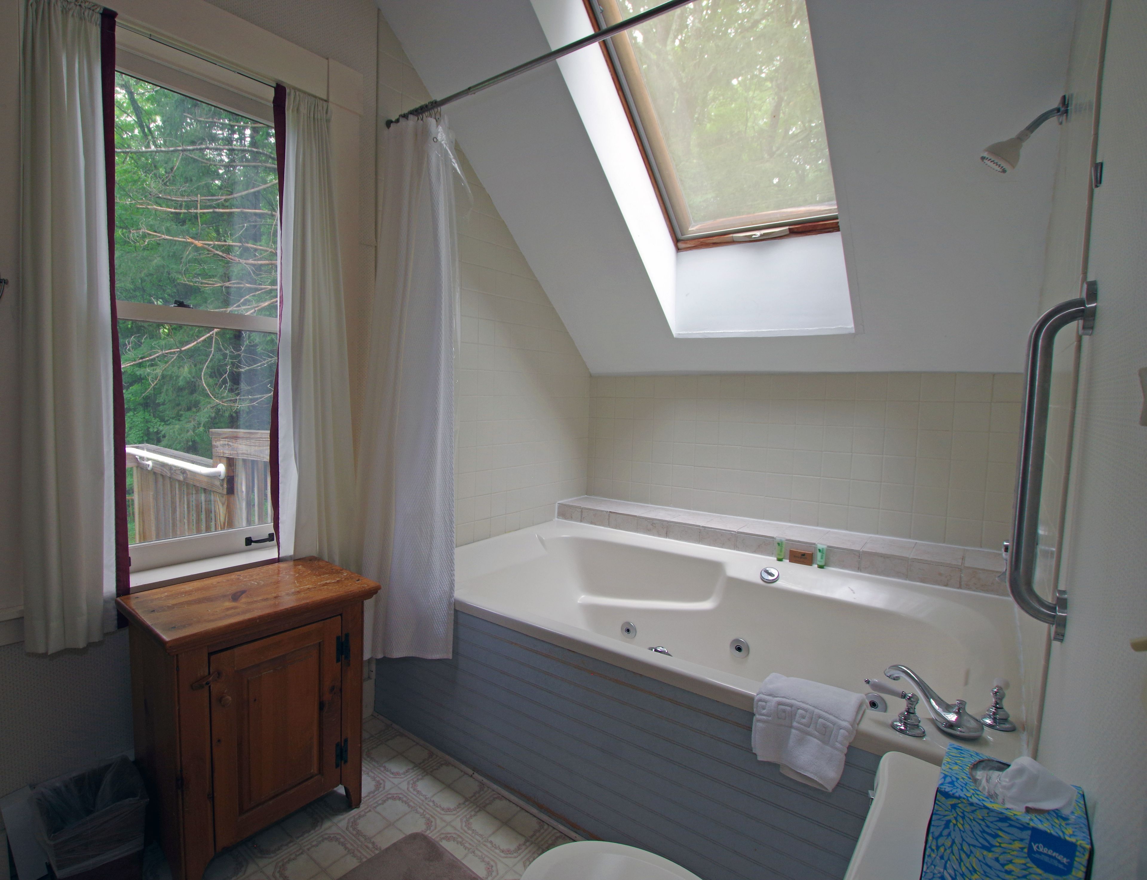A bathroom with a bathtub, a window with a view of trees, a wooden cabinet, and a shower area.