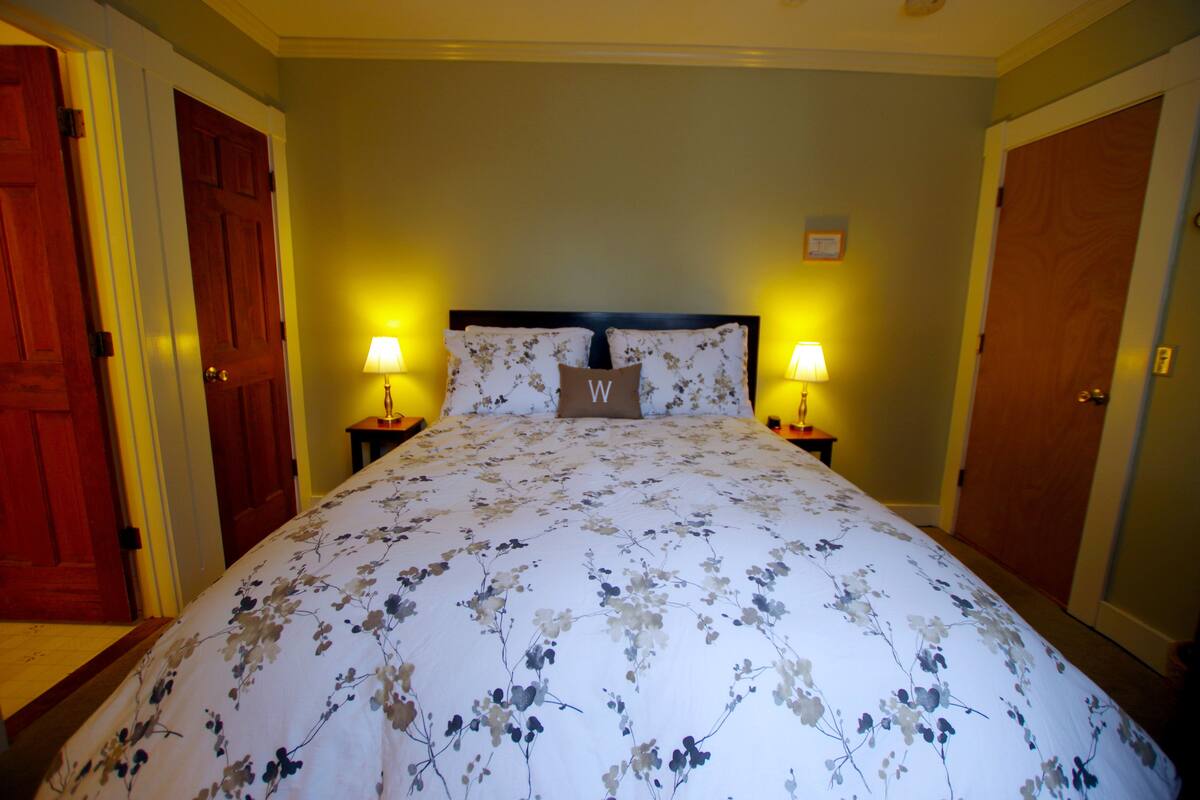 Une chambre d’hôtel avec une couvre-lit à motifs floraux, deux lampes de chevet, une tête de lit ornée d’un coussin en forme de « W », et des portes en bois.