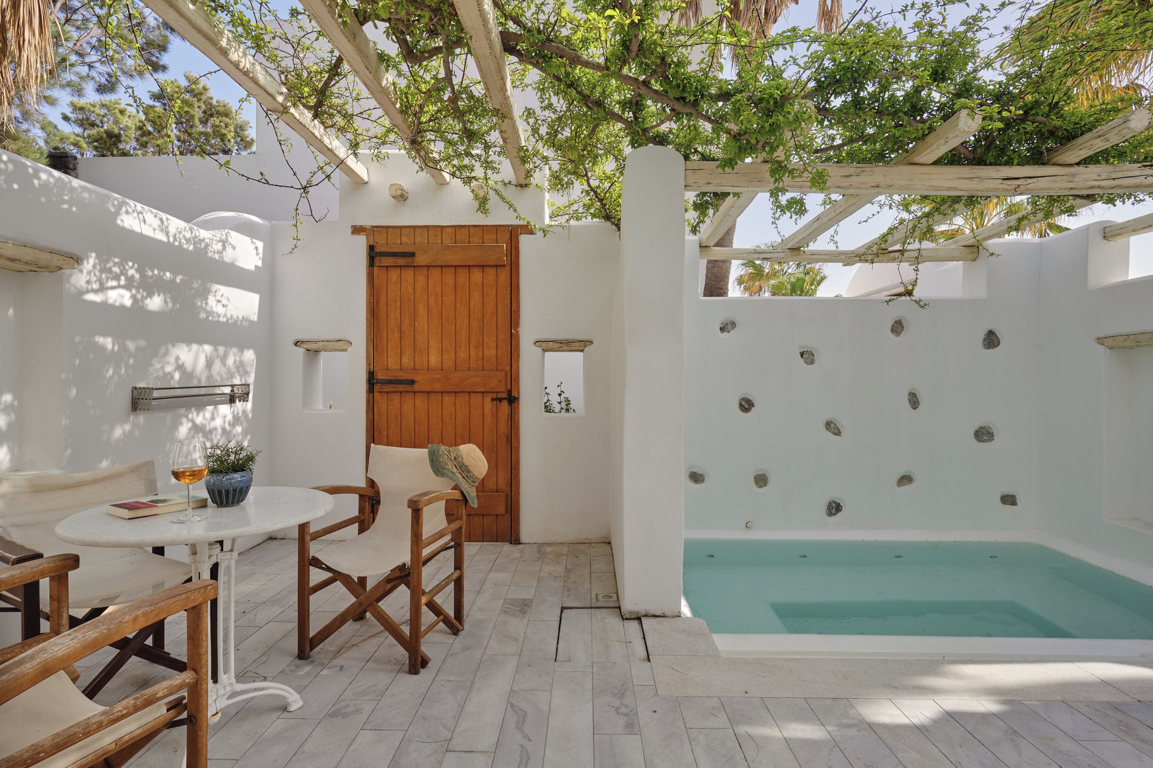 A white-walled room with a wooden door, a small round table with a glass, a chair, and a wooden bench.