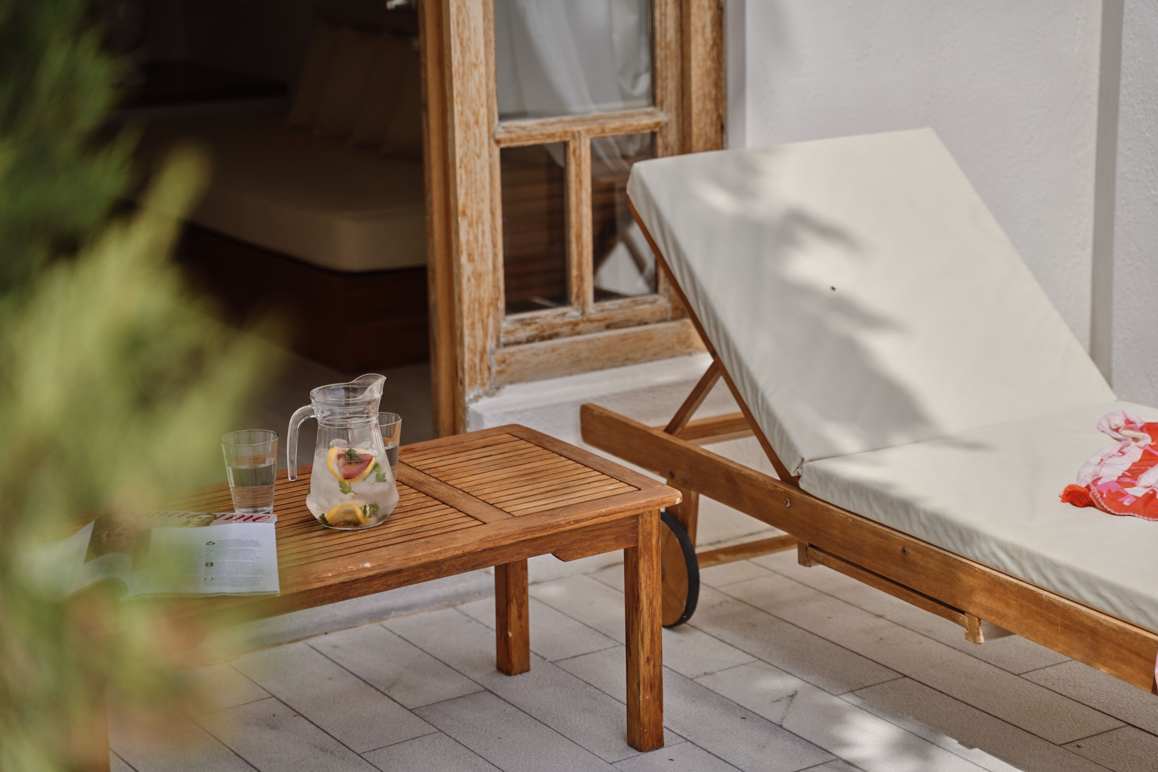 Una mesa de madera con una jarrón y vasos, una silla de madera con un cojín blanco, y una ventana con marco de madera.