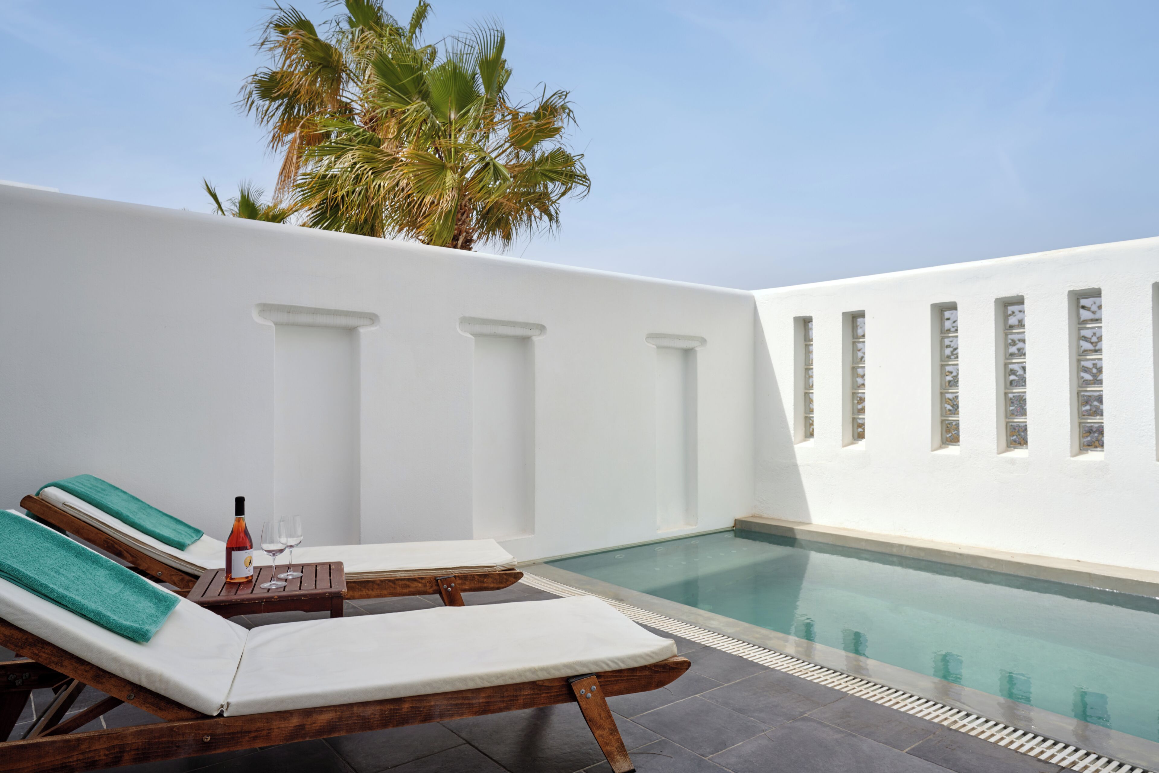 A poolside area with lounge chairs, a small table with a bottle and glasses, and a palm tree in the background.