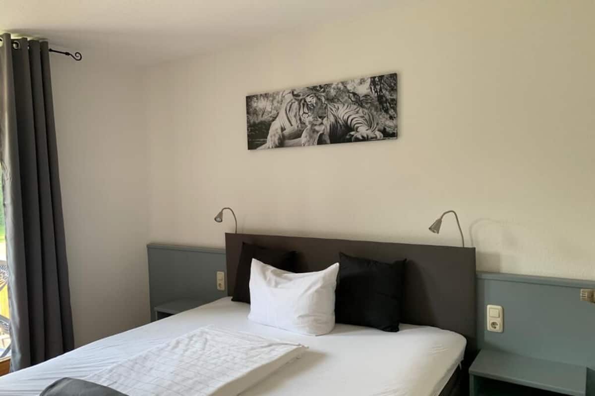 A hotel room with a bed, a headboard, a nightstand, a wall-mounted lamp, and a painting of a tiger.