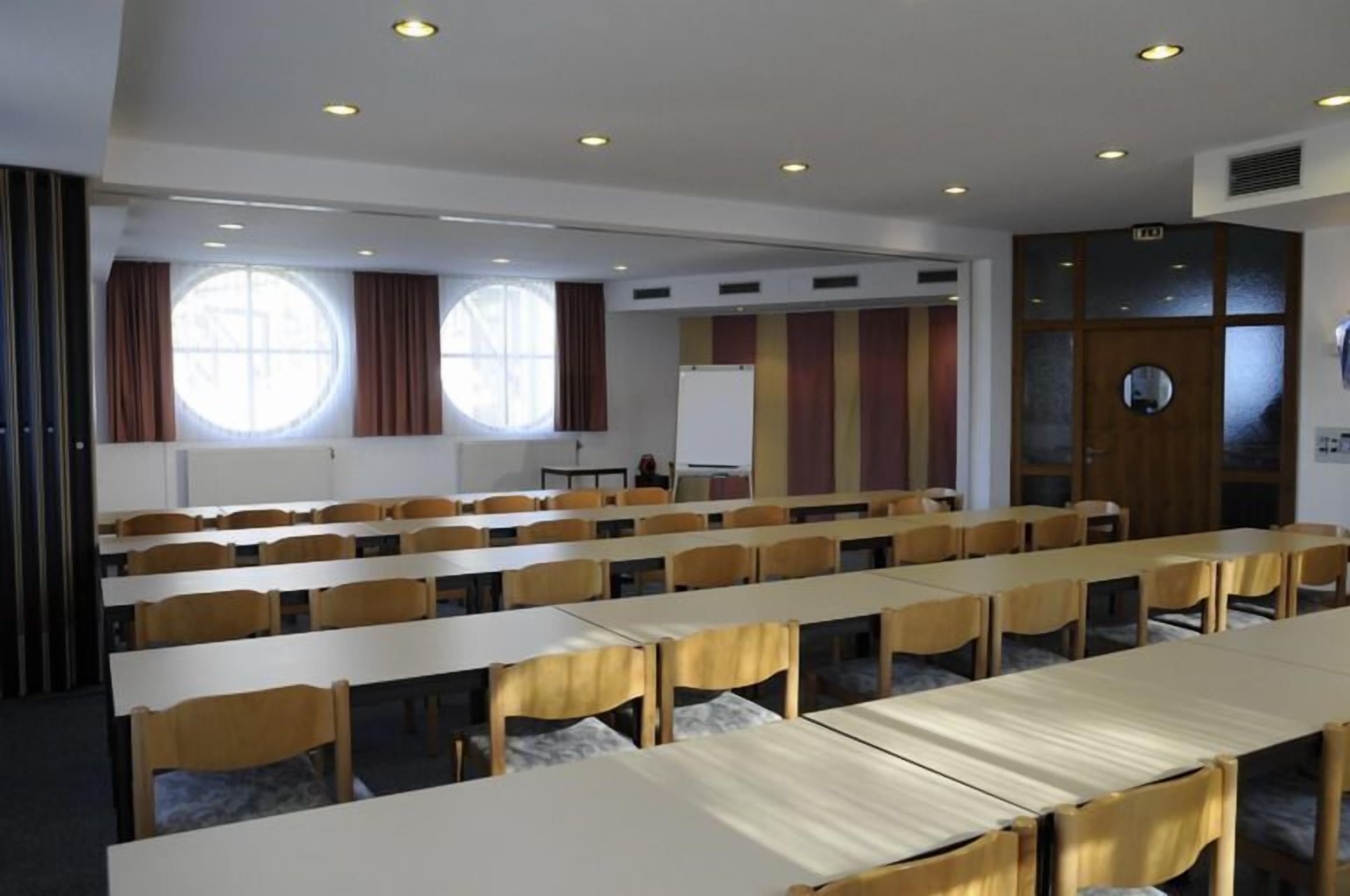 A conference room with rows of tables and chairs, large windows with curtains, and a whiteboard.