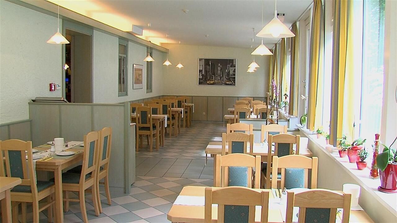 Ein Restaurant mit Holztischen und -stühlen, Fliesenboden, Pendelleuchten und einem Fenster mit Blumentöpfen.