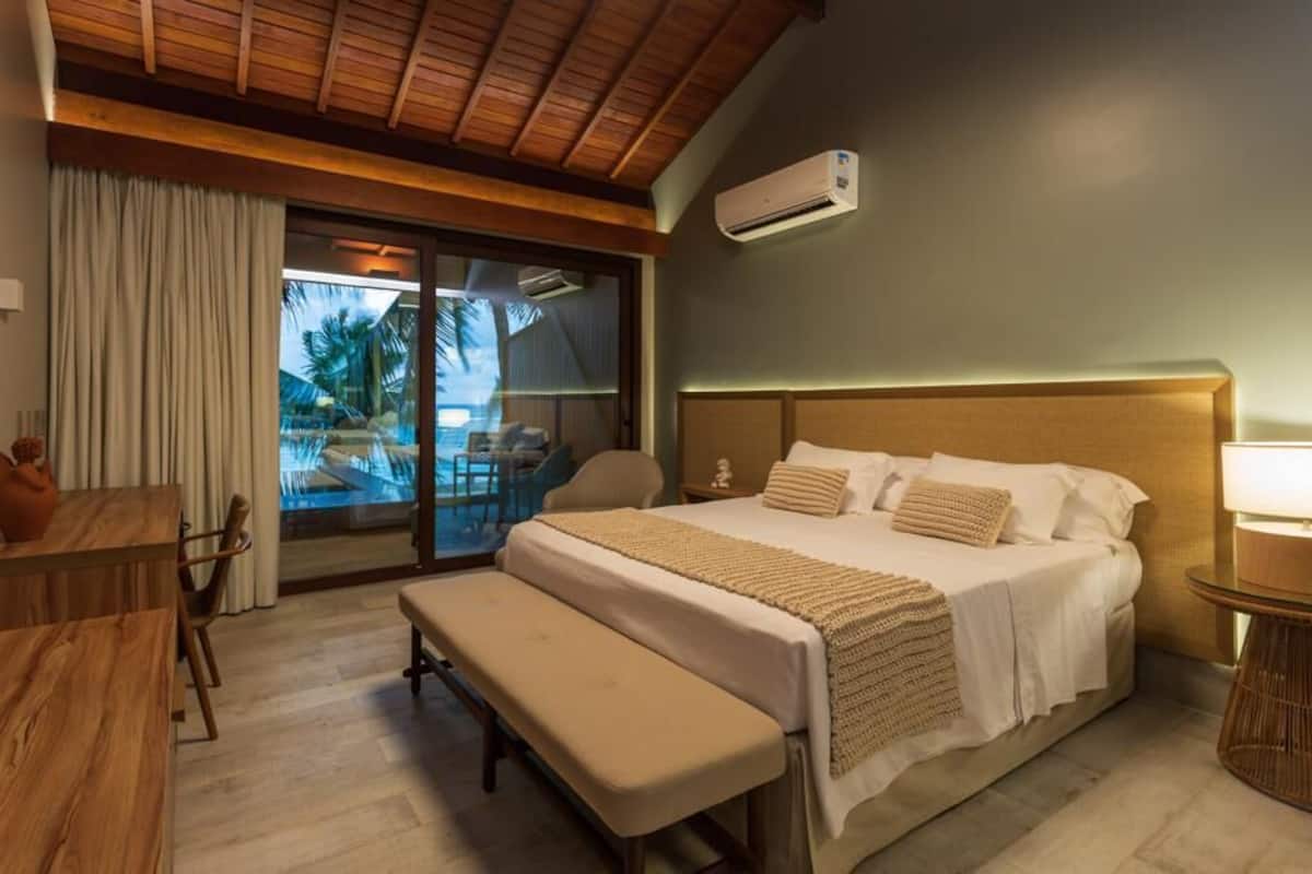 A hotel room with a bed, a bench, a table, and a view of a pool and palm trees.