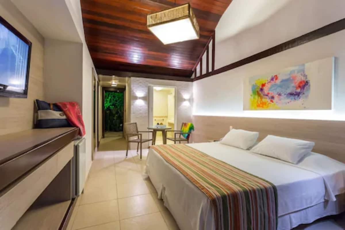 A hotel room with a bed, a desk, a chair, a TV, and a painting on the wall.