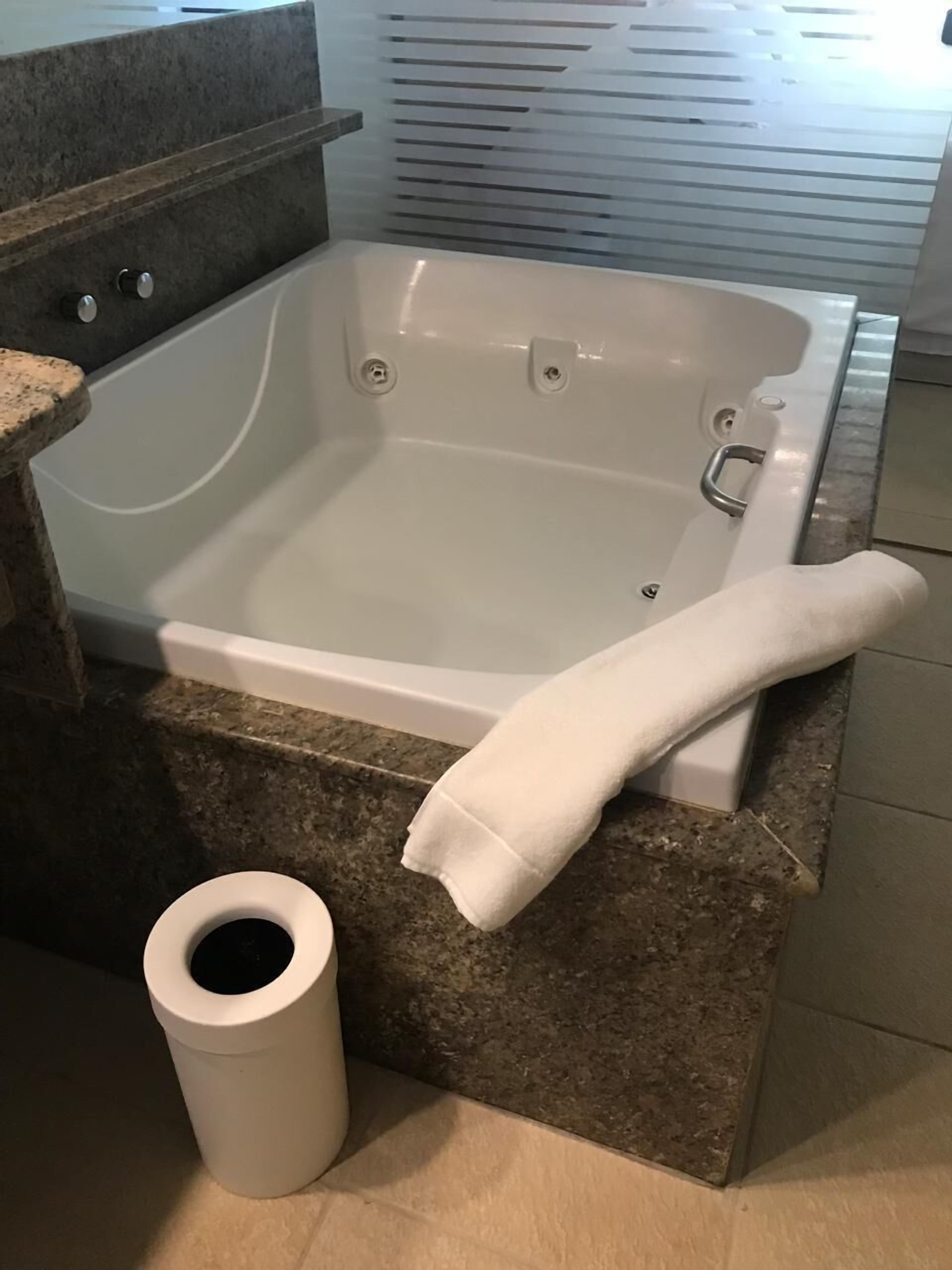 A bathroom with a white bathtub, a granite countertop, a white towel, and a trash can.