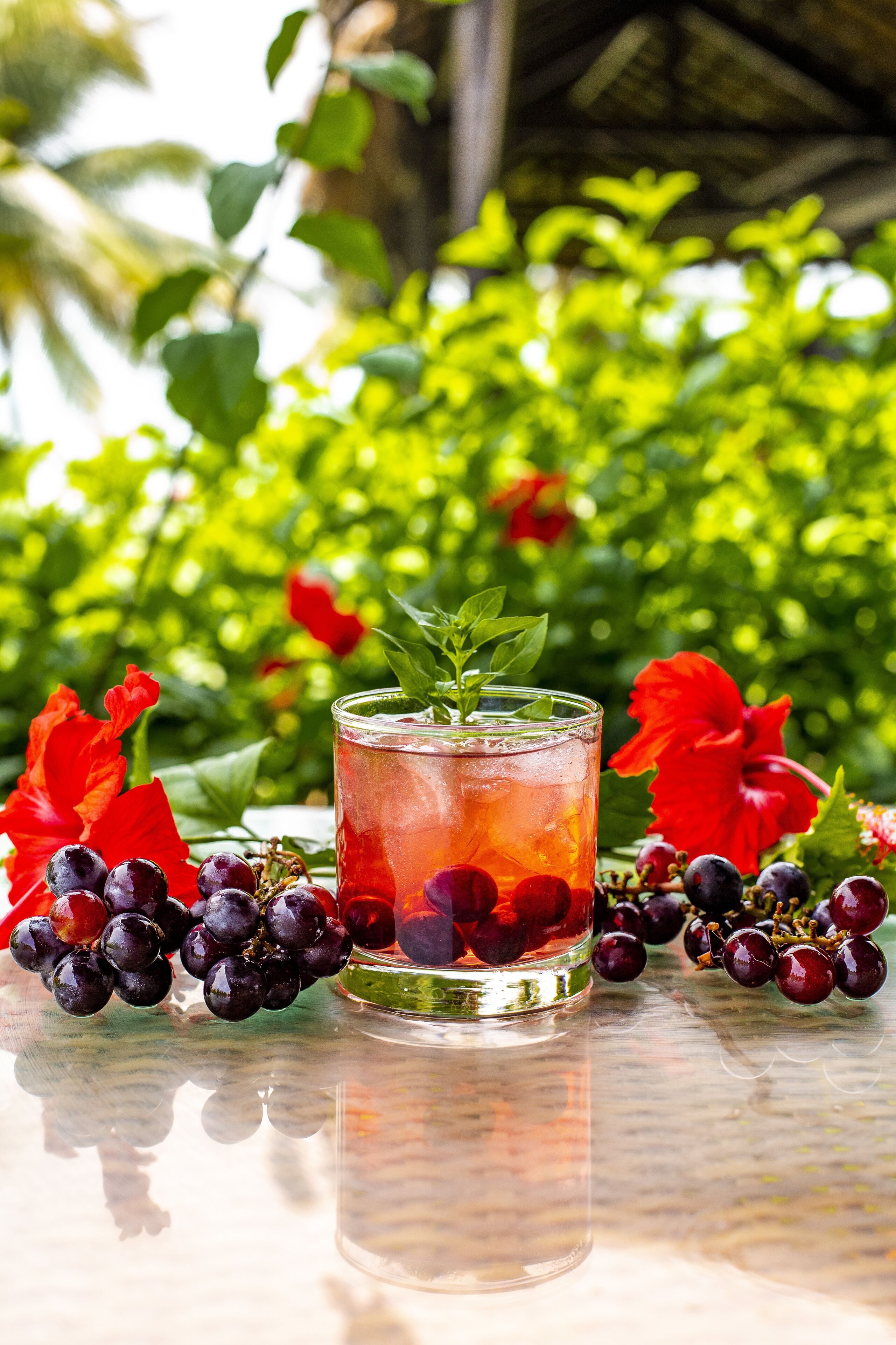 A glass of iced beverage with a mint garnish, surrounded by red hibiscus flowers and clusters of dark grapes on a reflective surface.