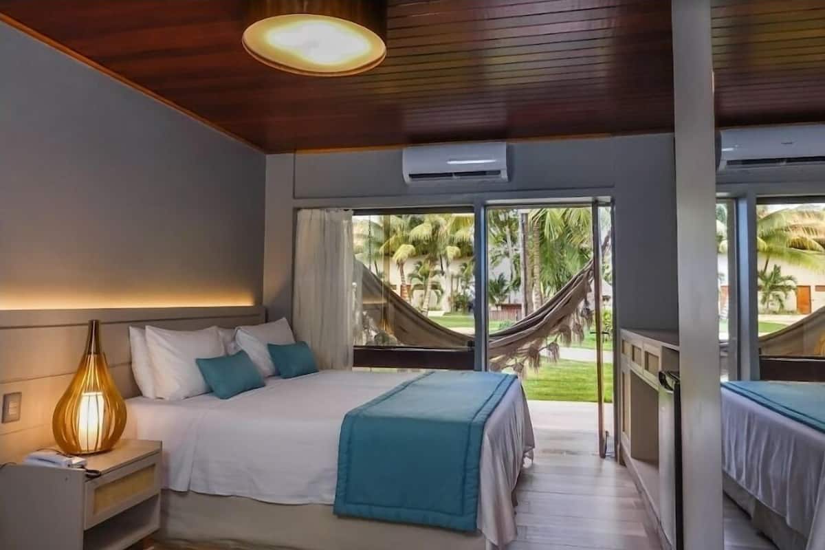 A bedroom with a bed, bedside lamp, and a view of a hammock and palm trees outside.