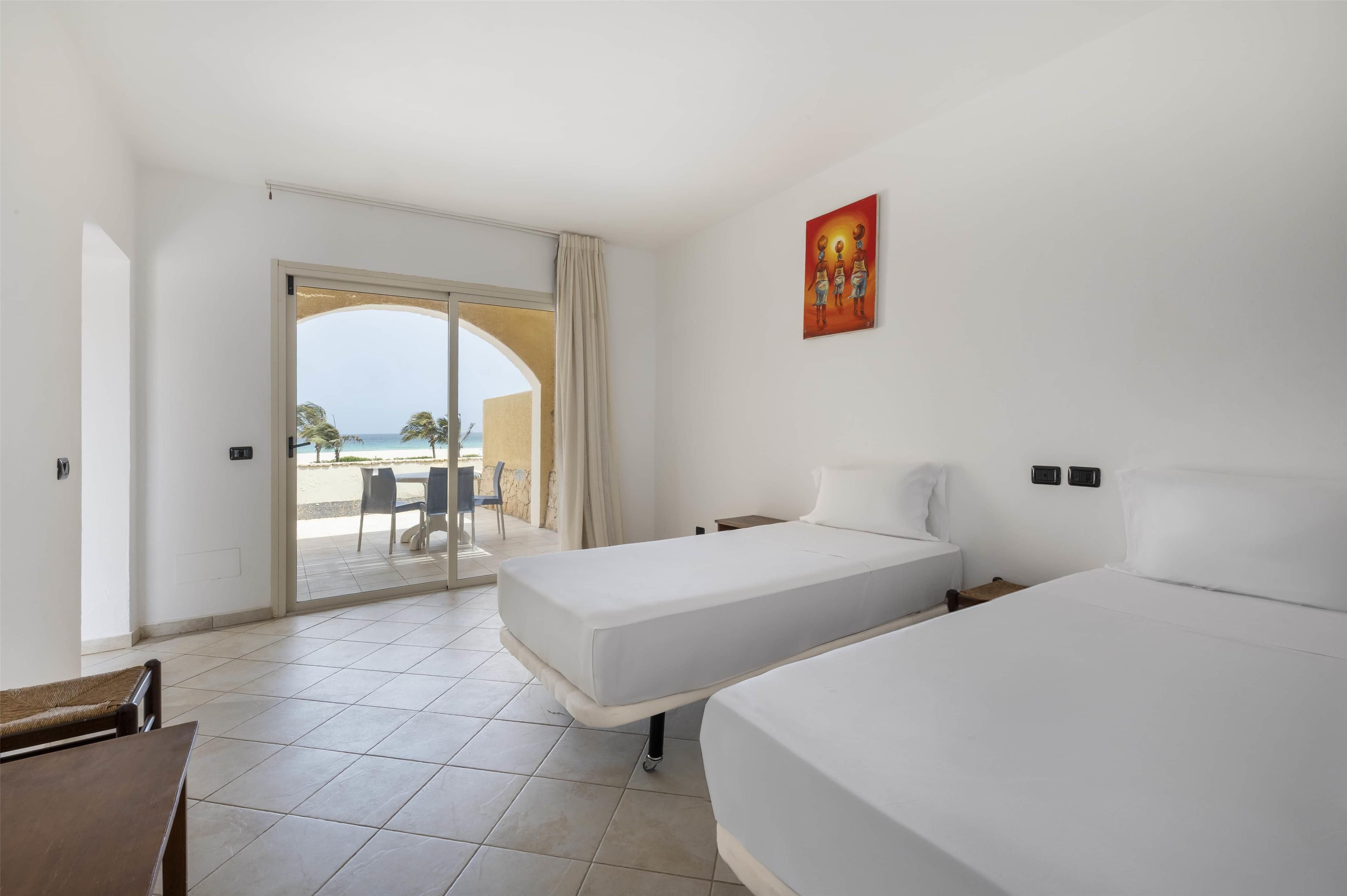 A hotel room with two beds, a view of the beach, and a dining area with chairs.