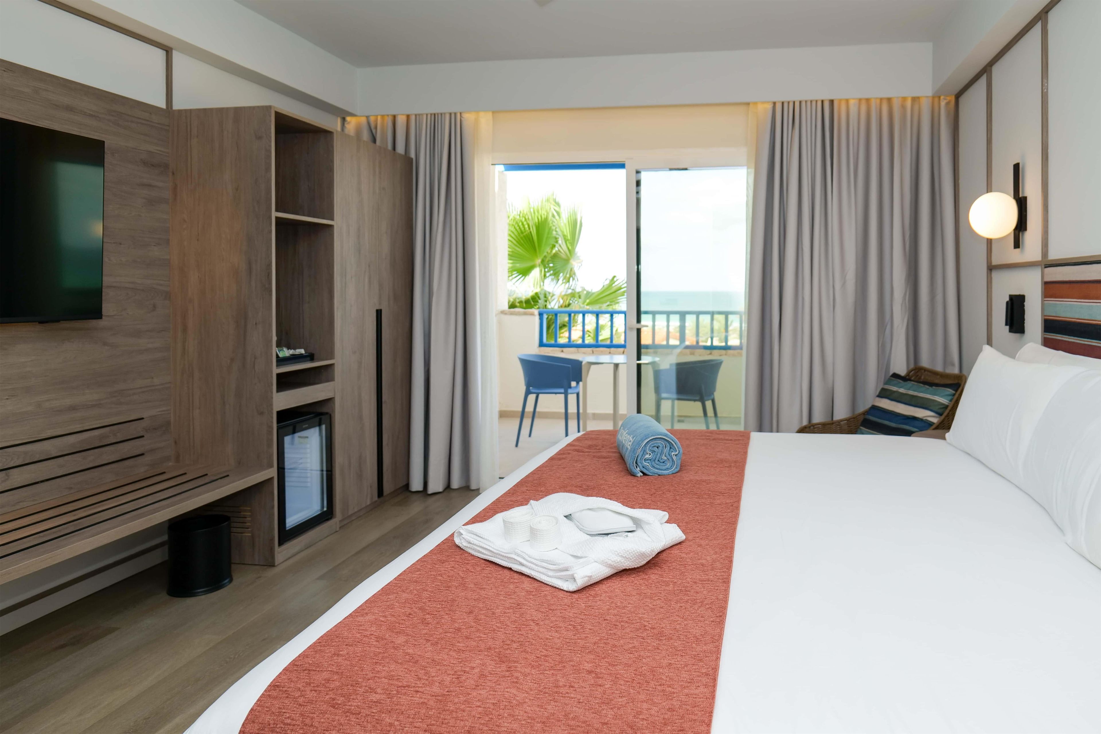 A hotel room with a bed, a TV, a minibar, a balcony with a view, and a dining area.