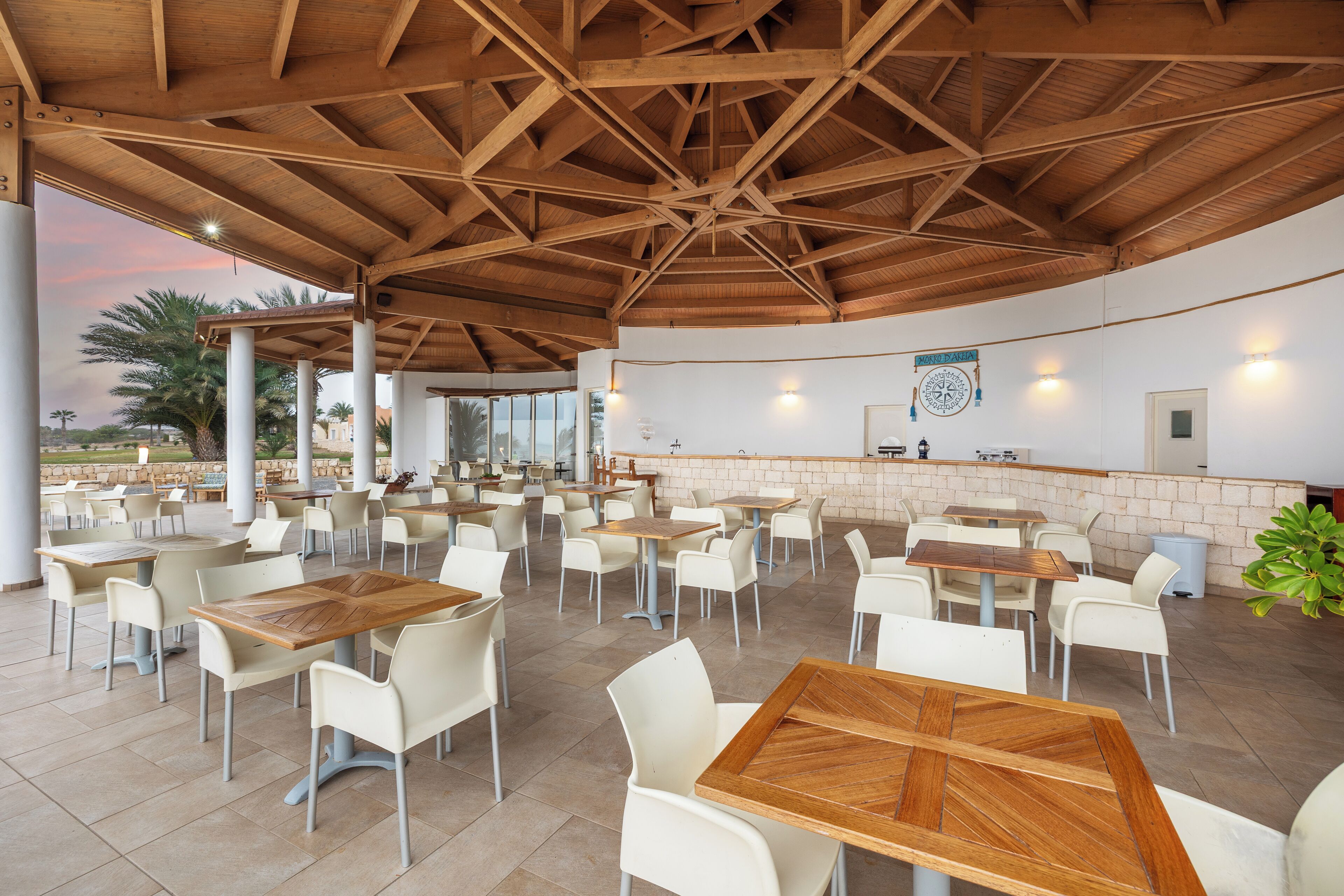 An outdoor café with wooden tables and white chairs, a brick wall with a sign, and a view of palm trees.