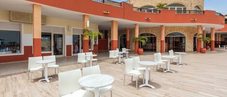 A spacious outdoor area with multiple white tables and chairs, a stone building with arches, and potted plants.