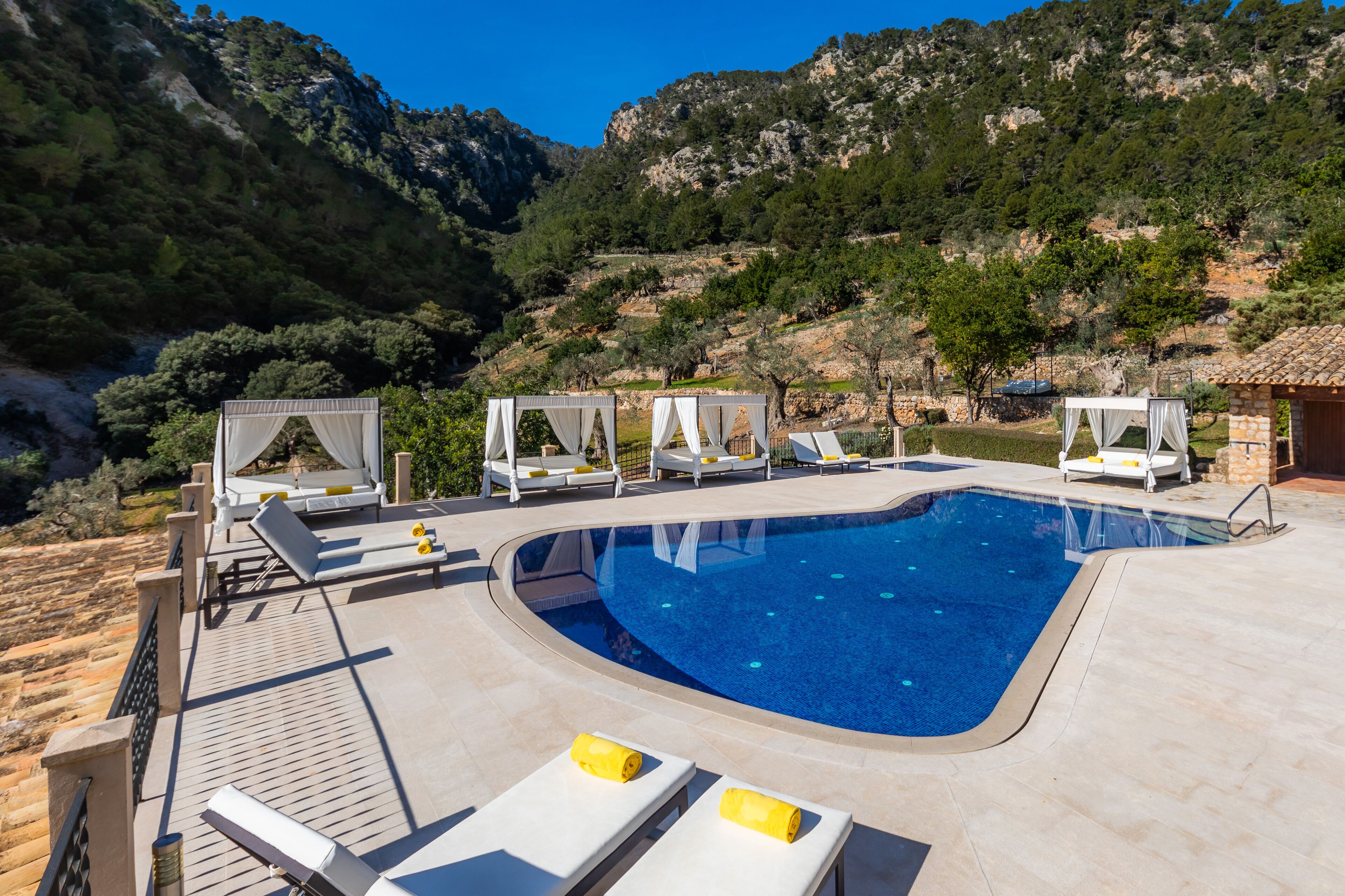 A swimming pool with sun loungers and cabanas, surrounded by a mountainous landscape.