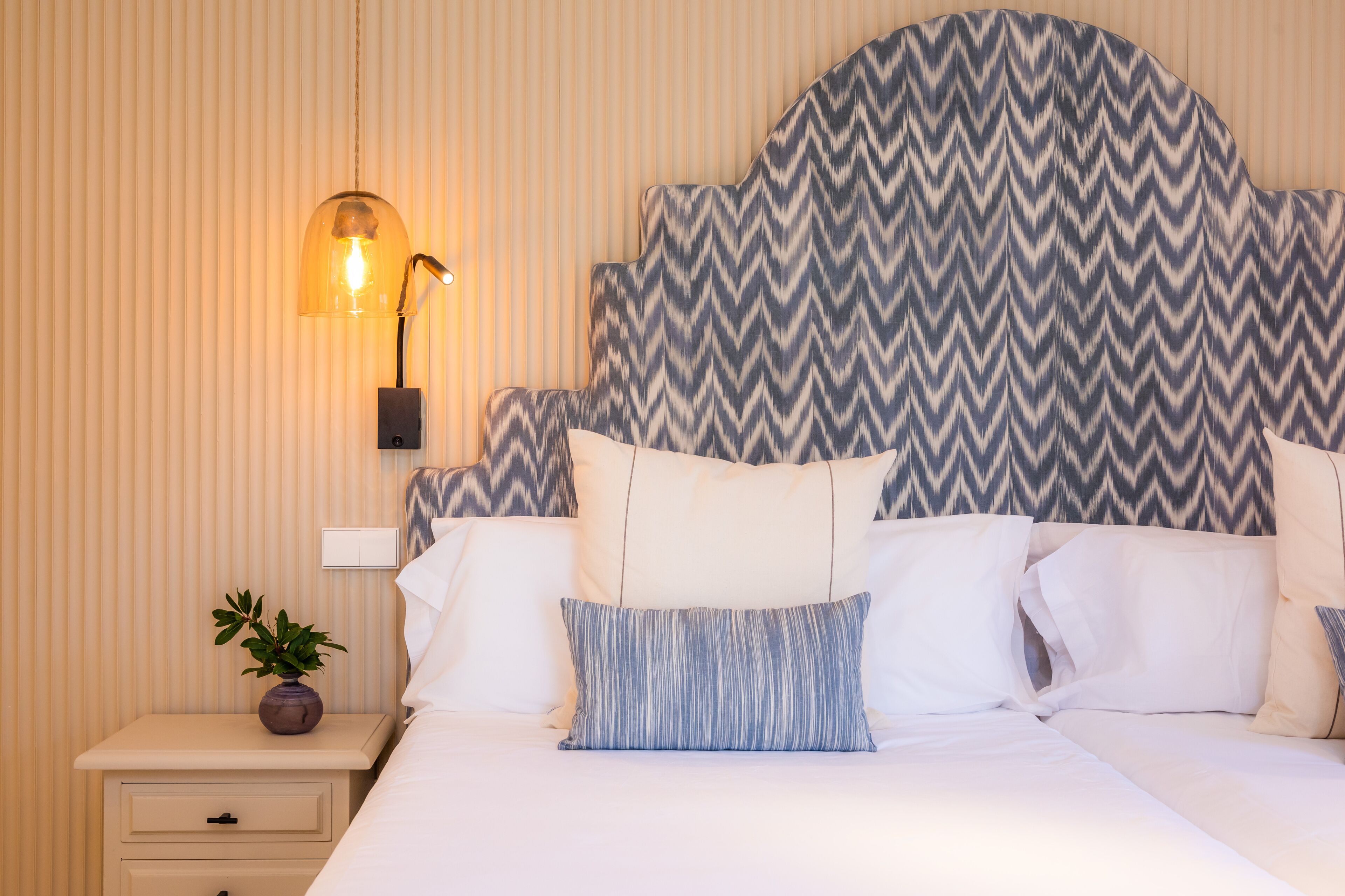 Una habitación de hotel con cama, almohadas, mesita de noche con una planta y una lámpara de pared.