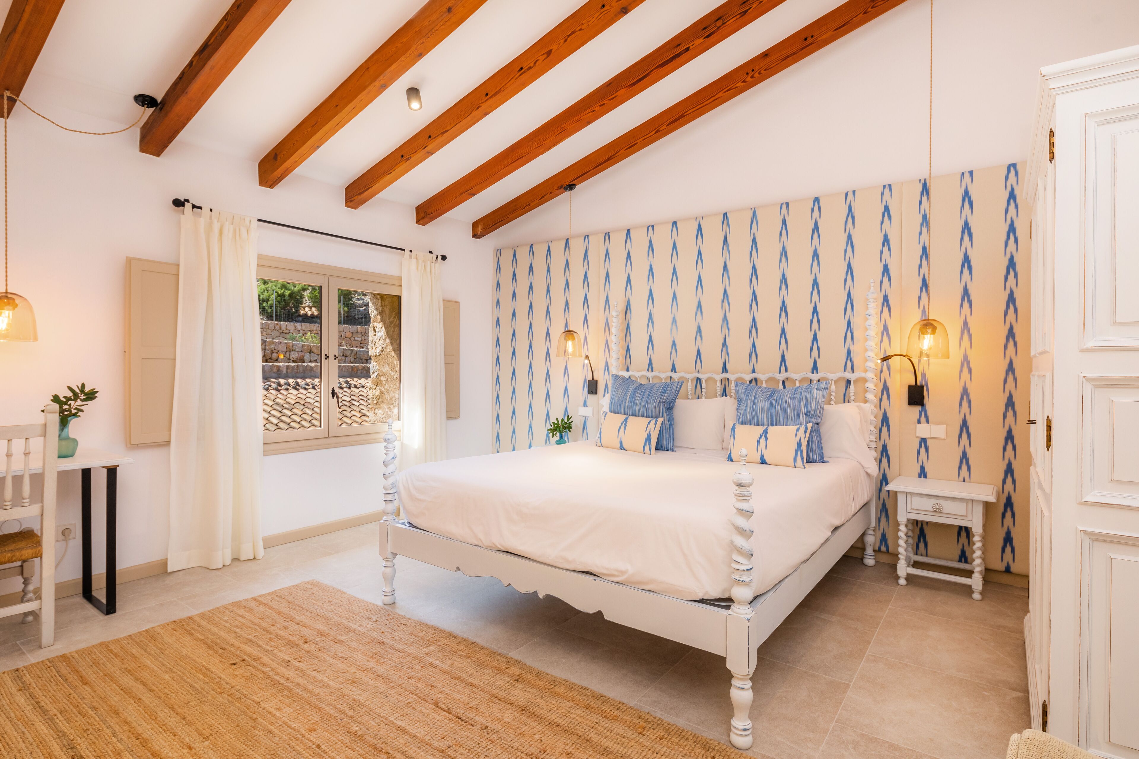 A bedroom with a white bed, wooden beams, patterned wallpaper, and a small bedside table with a lamp.