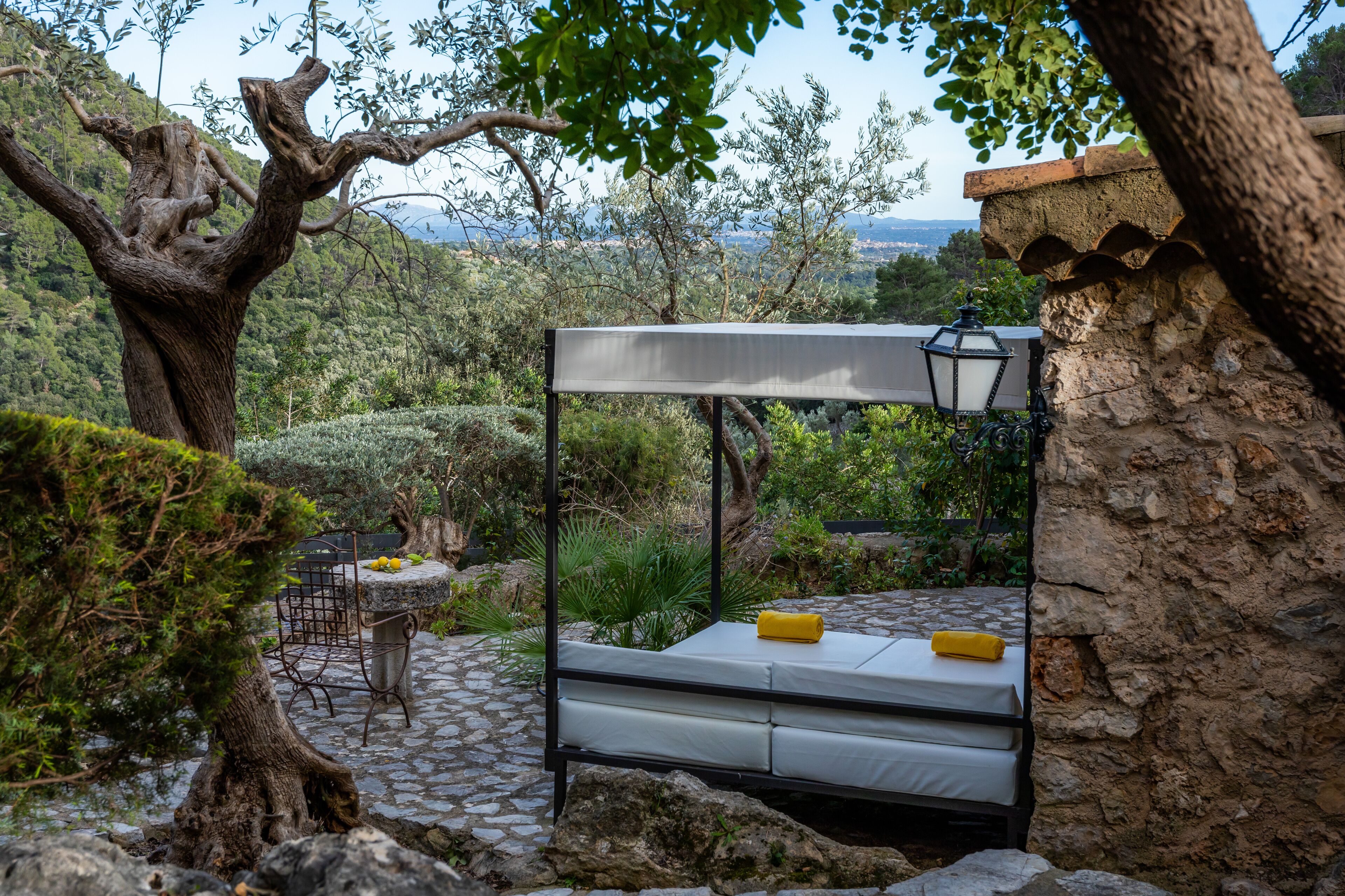 Un dormitorio rústico al aire libre con estructura de cama metálica, una mesita con sillas y un farol.