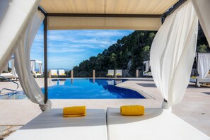 Zona junto a la piscina con toldos blancos, dos toallas amarillas y vistas a una piscina azul cristalina y una ladera boscosa.