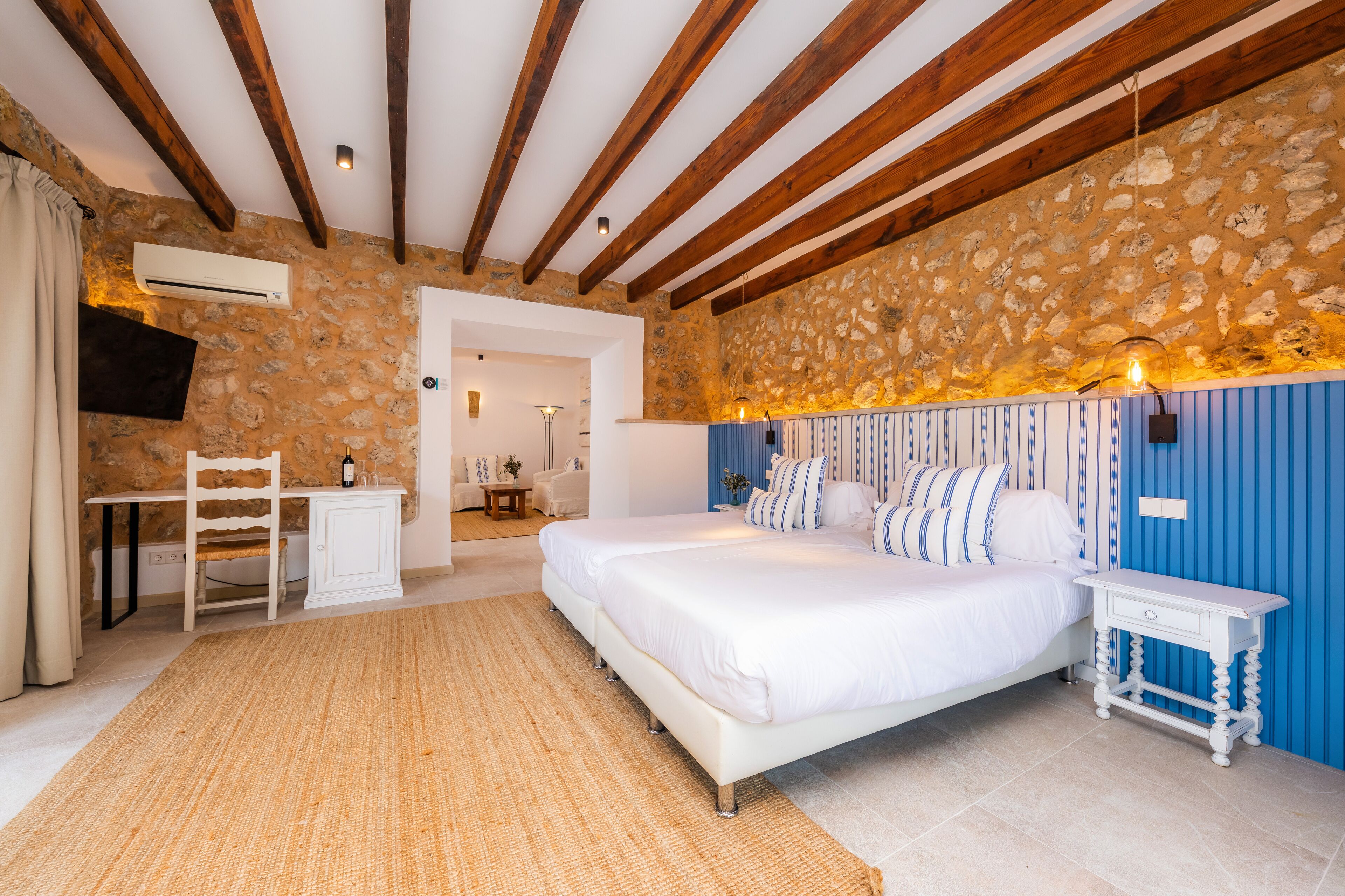 Un dormitorio con pared de piedra, vigas de madera, una cama, una mesita de noche, un escritorio y una silla.