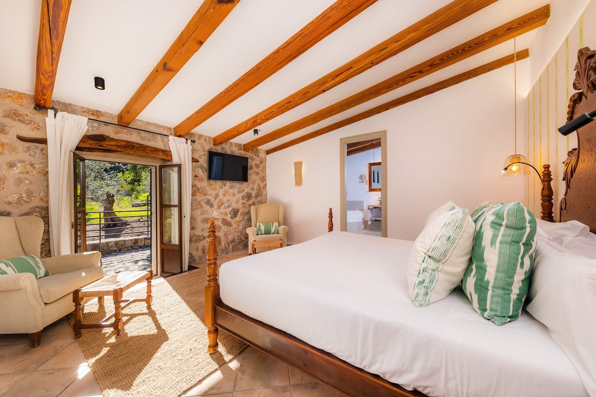 Ein Schlafzimmer mit einem hölzernen Bett, einer Steinwand, einer Holzbalkendecke, einem Fernseher und Blick ins Freie.