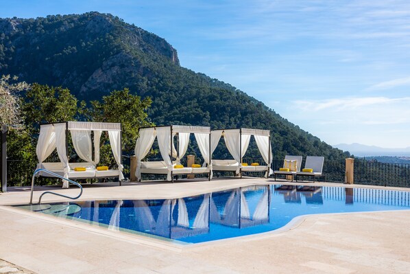 Un área de piscina con cuatro sillones blancos cubiertos por toldos, una piscina azul cristalina y un paisaje montañoso de fondo.