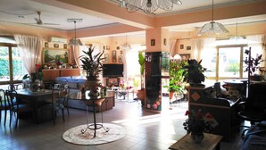 A spacious living area with a dining table, chairs, a sofa, and a TV. There are multiple potted plants and a ceiling fan. - Hotel Eliseo (Giardini Naxos)