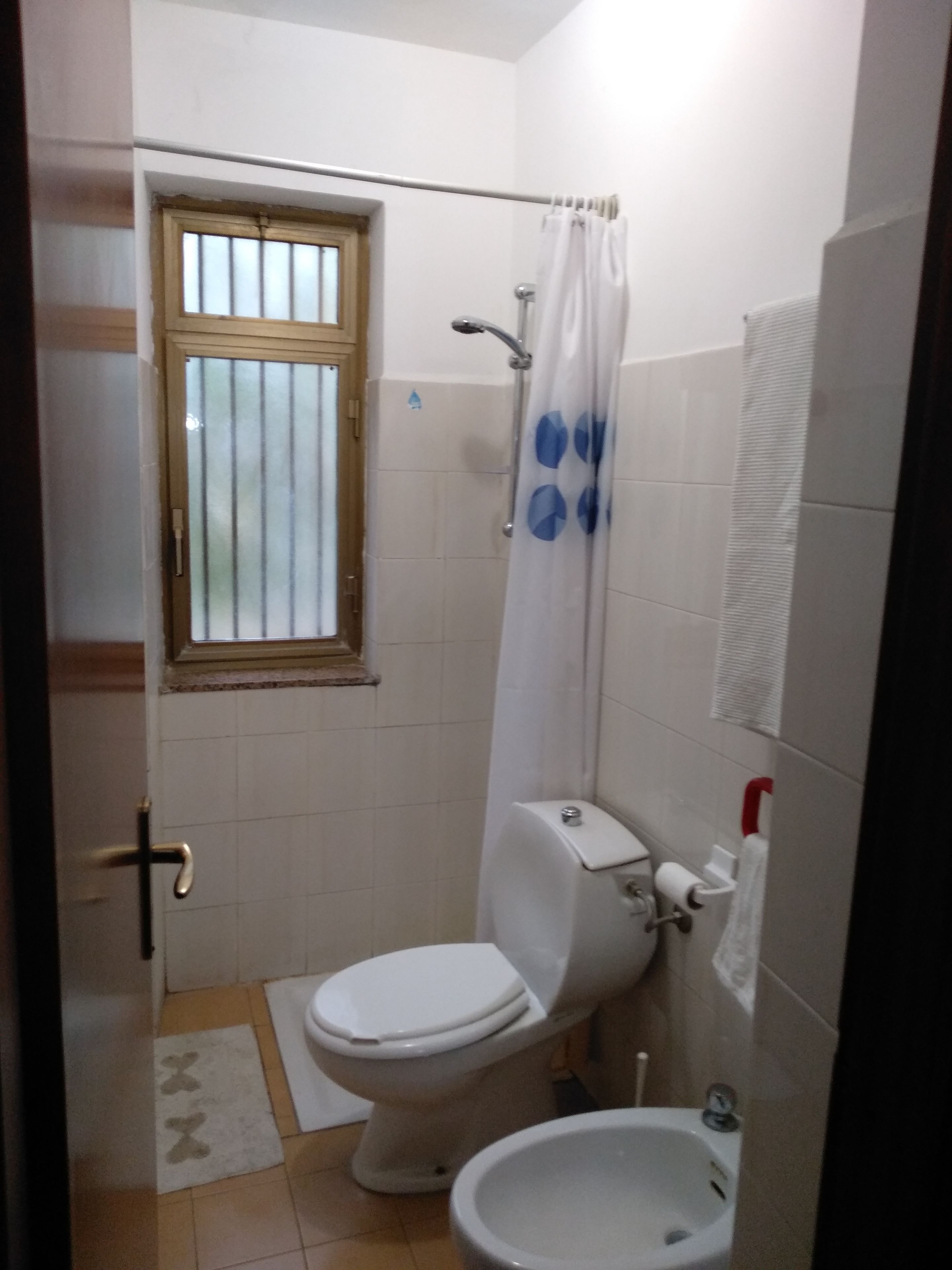 Une petite salle de bain compacte, équipée de toilettes, d’un bidet et d’une fenêtre avec des barreaux.