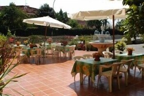 An outdoor patio with tables and chairs, some covered by umbrellas, and a fountain in the background. - Hotel Eliseo (Giardini Naxos)