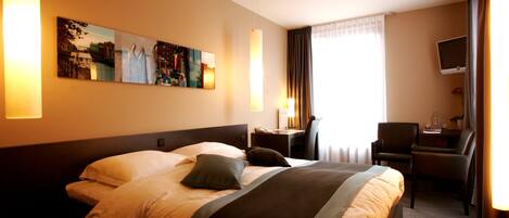 A hotel room with a large bed, a desk, a chair, a TV, and artwork on the wall.